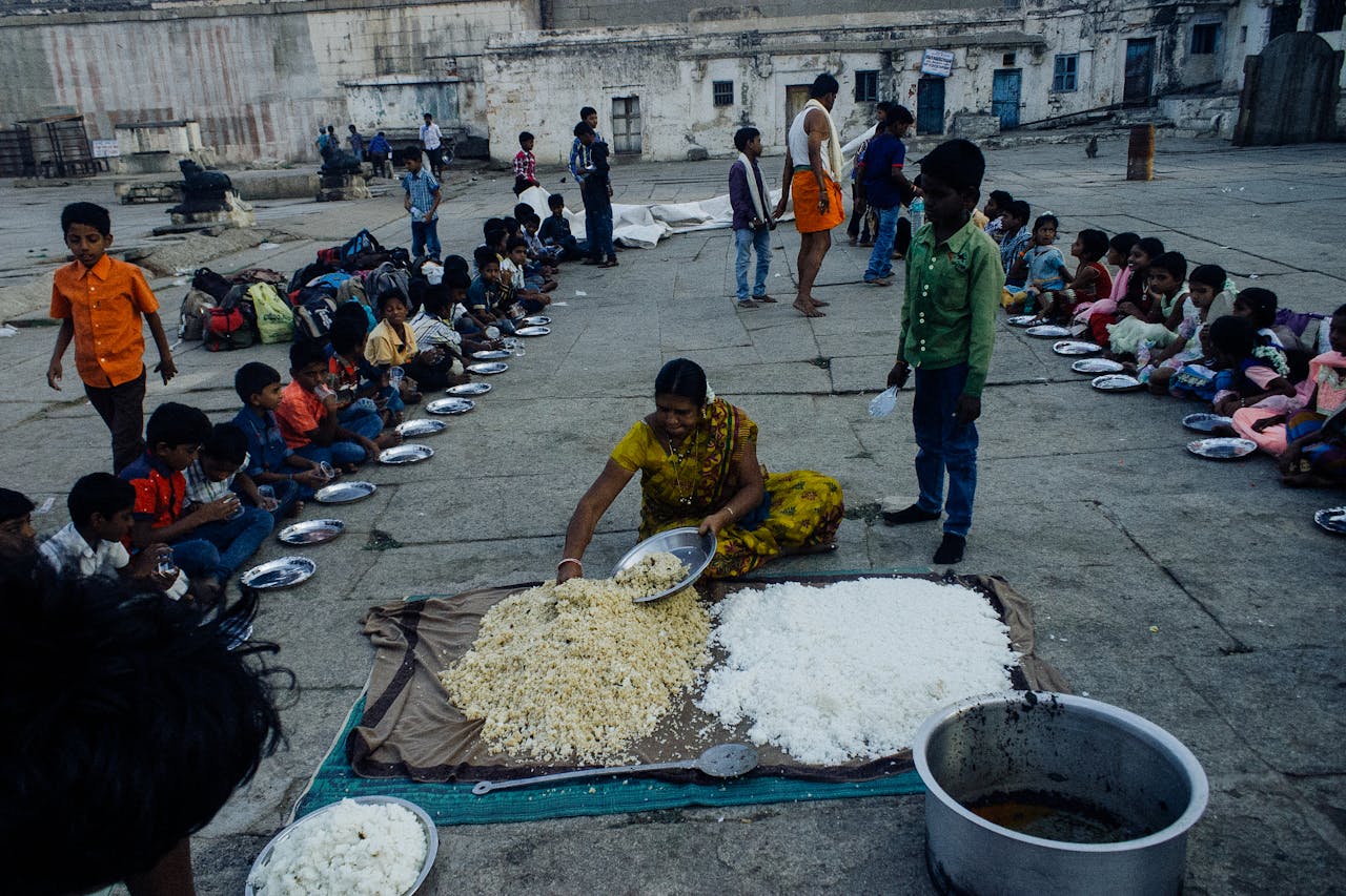 services-01 From above of old Indian woman serving food on plates while sitting on ground surrounded with kids in poor area
